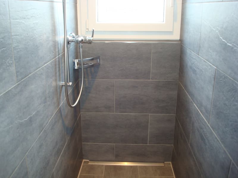Shower area with grey tiled walls and floor, chrome faucet and shower handle, small window above.