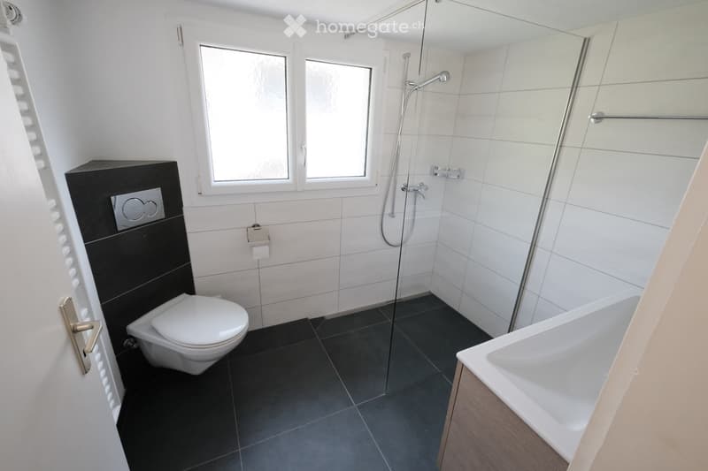 Modern bathroom with white tiles, black accents, a toilet, bathtub, shower, and a sink with a mirror above it.