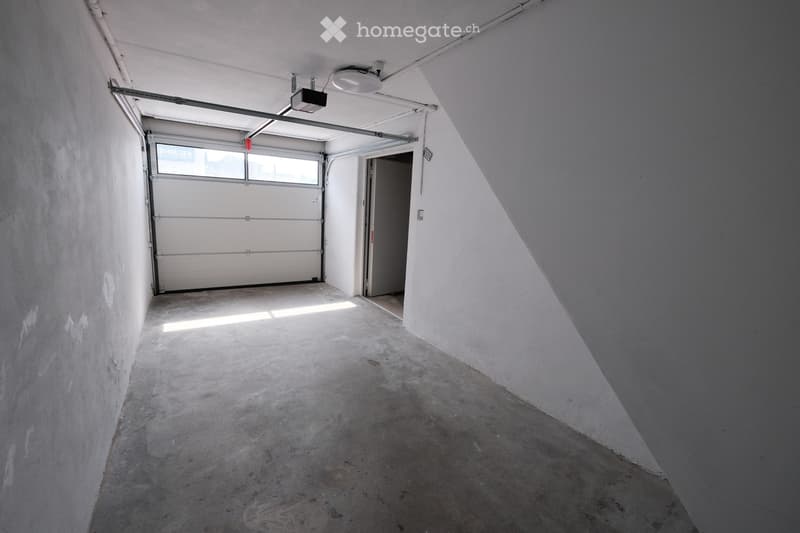 Empty garage with a concrete floor, white walls, and a large overhead door. There is a small doorway leading to another area.