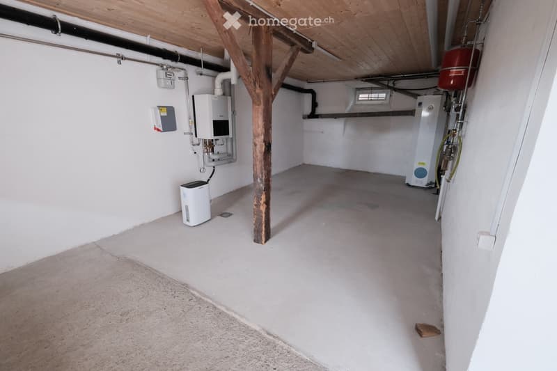 Empty basement room with white walls, concrete floor, exposed wooden beam, and several utility appliances including water heaters and electrical panels.