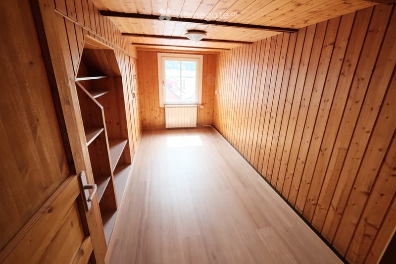Empty wooden hallway with slanted ceilings, wooden walls, and a window at the end. The floor is light wood, and there are built-in shelves on one side.