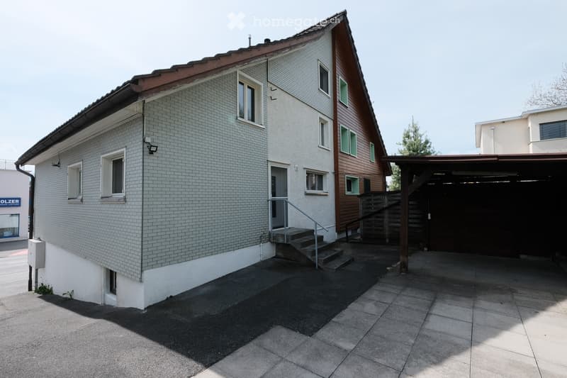 Two-story house with a tiled roof, white brick facade, and wooden siding on one side. Features include a staircase leading to the entrance, a covered garage area, and a paved patio.