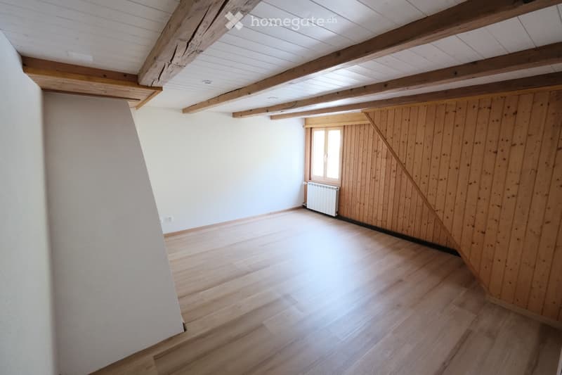 Empty room with wooden flooring, exposed wooden beams on the ceiling, white walls, and a single window with wooden shutters.