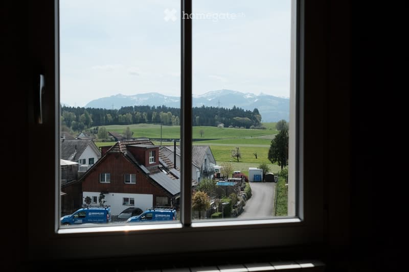 View from a window showing a rural village with houses, green fields, and distant mountains.
