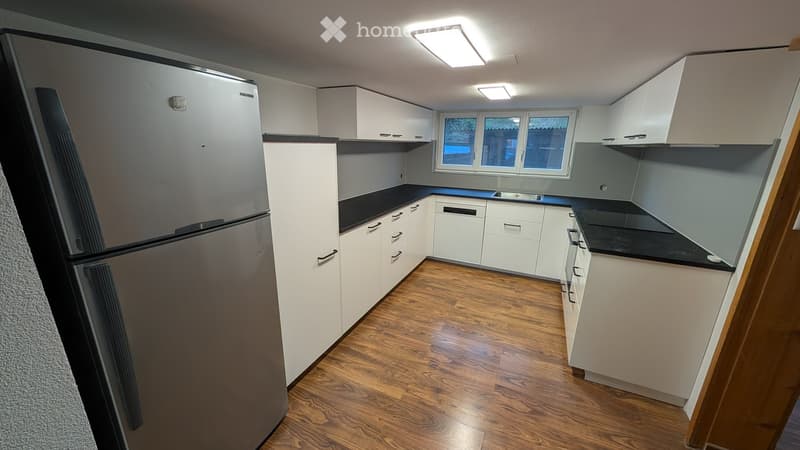 Modern kitchen with white cabinets, black countertops, and a stainless steel refrigerator. The floor is made of wood, and there is a window above the sink.