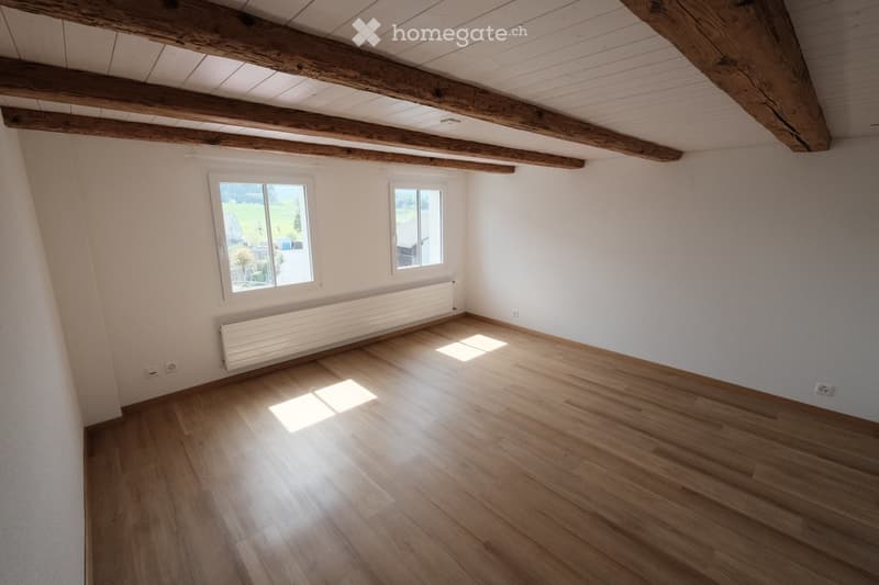Empty room with wooden flooring, white walls, exposed wooden beams on the ceiling, and two windows offering a view of the outdoors.