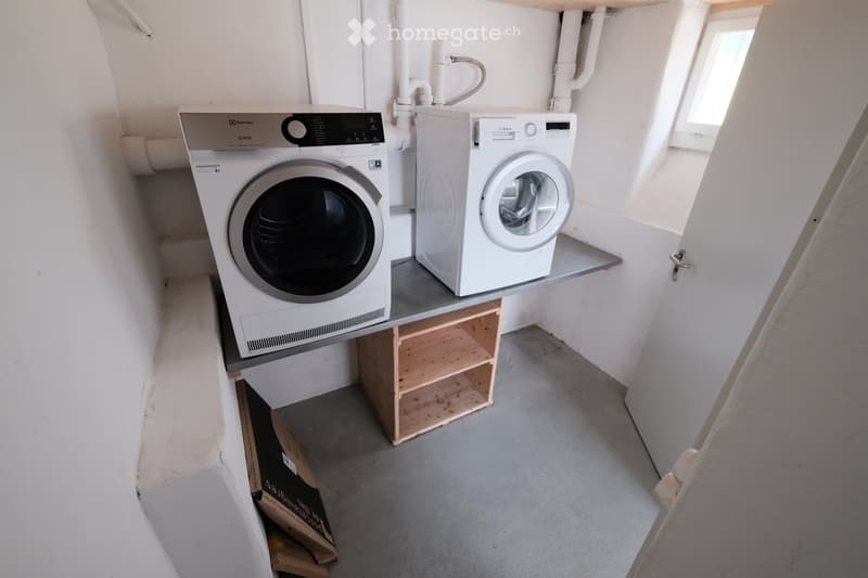 A small, well-lit room with a washing machine and dryer placed on a wooden shelf, white walls, and a grey concrete floor.