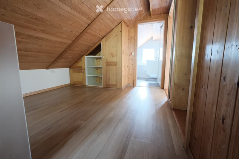 Empty attic space with wooden floors and slanted wooden ceilings, featuring built-in wooden shelving units and a doorway leading to another room.