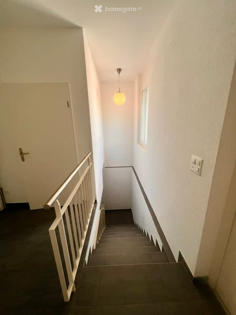 A narrow hallway with a staircase leading upwards, white walls, a single window, and a hanging light fixture.