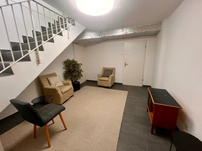 A small, modern living space with a cream-colored sofa, a wooden chair, a small table, a potted plant, and a cabinet. The room features a staircase leading upwards and a white door.