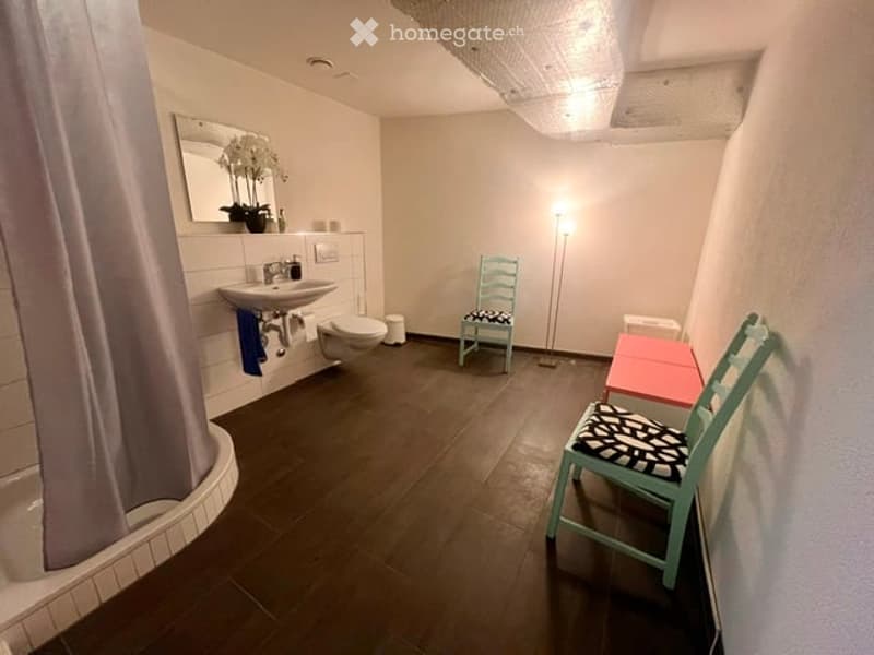 Modern bathroom with dark wooden flooring, white walls, bathtub, sink, toilet, and a small seating area with a chair and table.