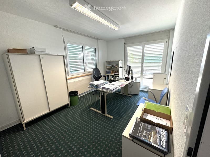 An empty office with a desk, chair, computer, and cabinet. The room has a carpeted floor, white walls, and two windows with blinds. There is a trash bin and a shelf with books and a monitor.