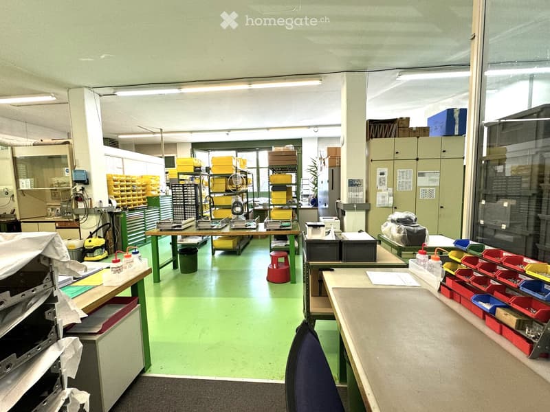 A workshop with green floor, multiple tables, shelves, and storage boxes, a red stool, and a blue chair.