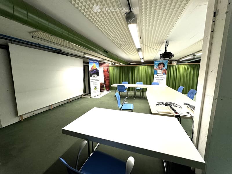 Empty room with white walls, green carpet, long white tables, blue chairs, whiteboard, green curtains, posters, projector