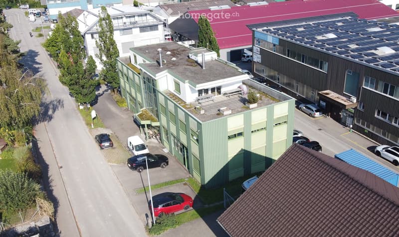 Aerial view of a building with multiple floors, green roof, and parking area with several cars parked.
