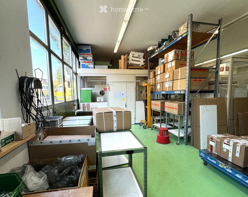 Warehouse with large windows, green floor, shelves with boxes, trolleys, and storage boxes