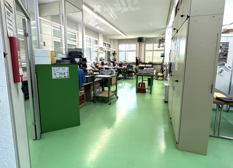 Industrial room with green floor, multiple desks, storage boxes, trolleys, and a fire extinguisher