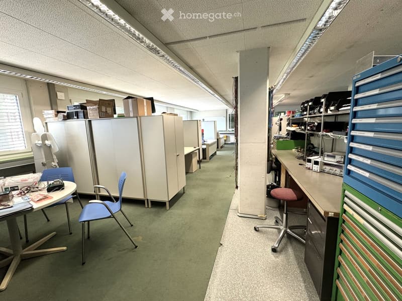 Office space with cubicles, tables, chairs, and storage boxes. Fluorescent ceiling lights, green carpet, and a window with blinds.