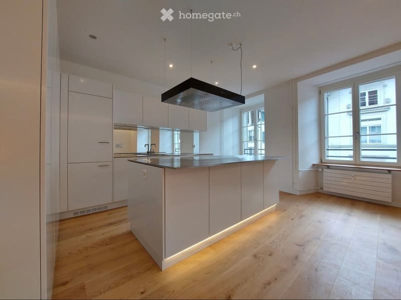 Modern kitchen with white cabinetry, a central island, and a range hood. The space features wooden flooring and large windows allowing natural light.