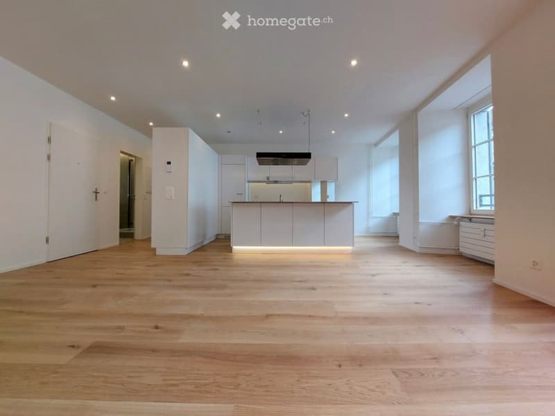 Open-concept unfurnished room with light wooden flooring, white walls, and built-in cabinetry. Features recessed ceiling lighting and large windows allowing natural light.