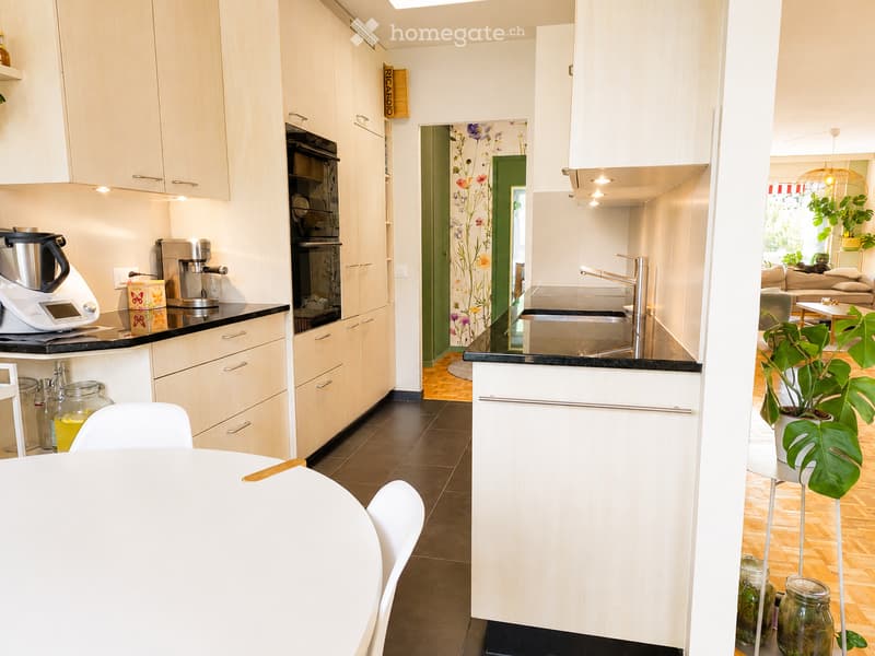Modern kitchen with white cabinets, black countertops, coffee maker, blender, sink, and a small dining area with a table and chairs. A plant is visible in the adjacent living space.