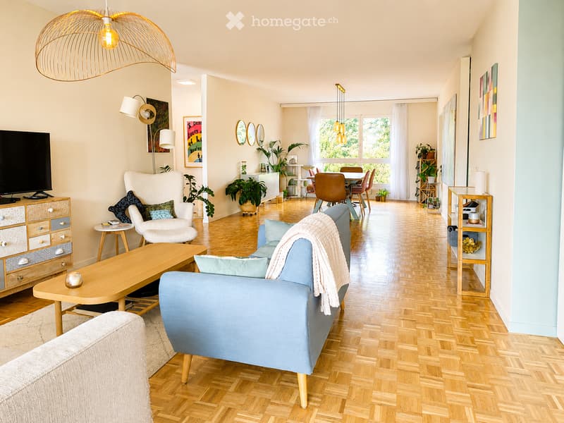 A modern living room with light wooden flooring, a blue sofa, a white armchair, a wooden coffee table, a TV on a cabinet, and a dining area with a table and chairs. The space is decorated with plants, artwork, and a large pendant light.