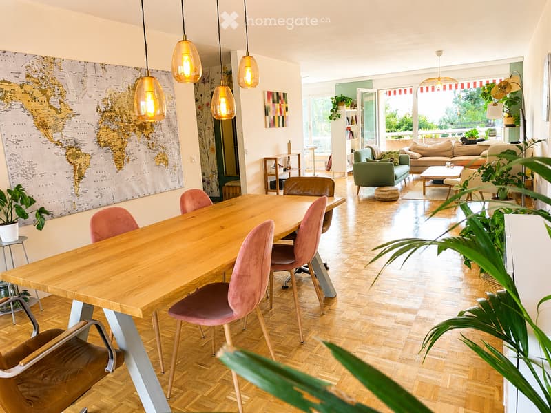 Open-plan living and dining area with wooden flooring, a large dining table with pink chairs, a green sofa, a coffee table, pendant lights, and plants.