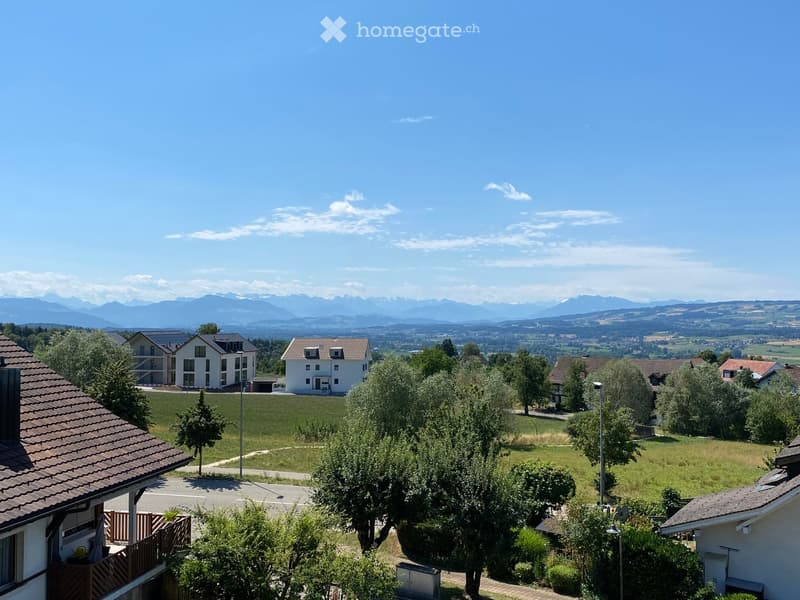 Residential area with multiple houses, green lawns, trees, and distant mountain range under a clear blue sky.