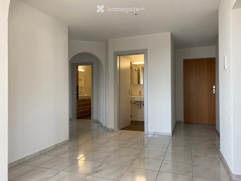 A clean, tiled hallway with white walls, featuring an arched doorway leading to a brightly lit bathroom and a closed wooden door on the right.