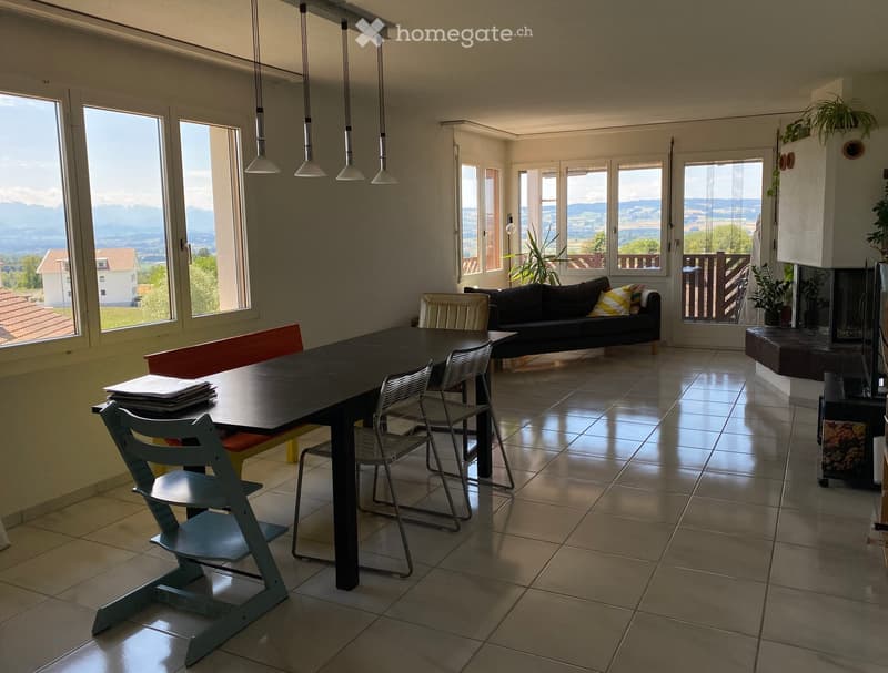 Bright living and dining area with large windows offering a scenic view, featuring a black sofa, a dining table with chairs, and pendant lights.
