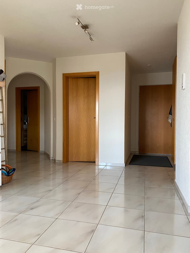 Empty hallway with tiled floor, wooden doors, and recessed lighting. Features an arched entryway and a small basket with items on the left side.