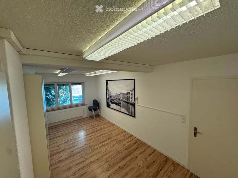 A small, unfurnished room with wooden flooring, white walls, a large window, a single chair, and a large picture frame on the wall.