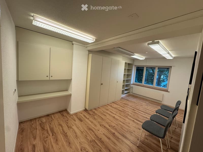 A small, unfurnished room with light wooden flooring, white walls, and built-in cabinets. There are four grey chairs arranged in a row, a window with a view of trees, and a shelf unit with open compartments.
