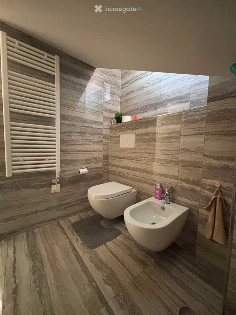 Modern bathroom with marble tiles, a toilet, a sink, a radiator, and a towel hanging on the wall.