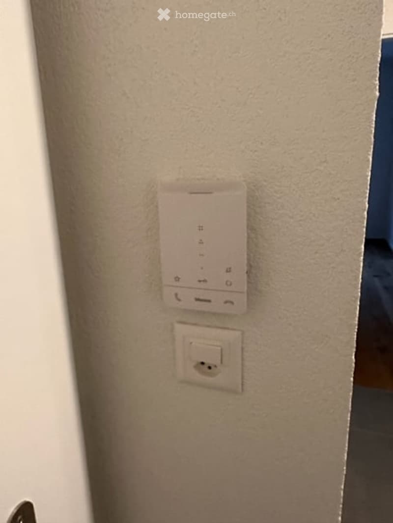 A close-up view of a wall showing a smart thermostat and an electrical outlet. The wall is painted a light beige color.