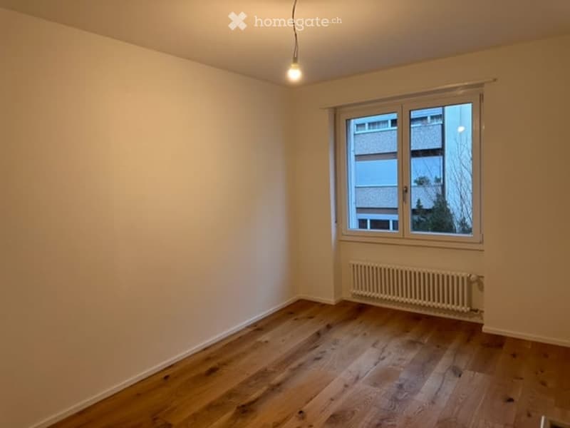 Empty room with wooden flooring, white walls, a single window with white frame, and a radiator below the window. No furniture or equipment present.