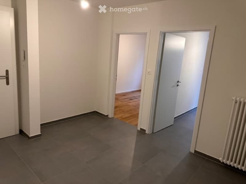 Empty room with tiled floor, wooden flooring visible through an open doorway, white walls, and a radiator on the right side.