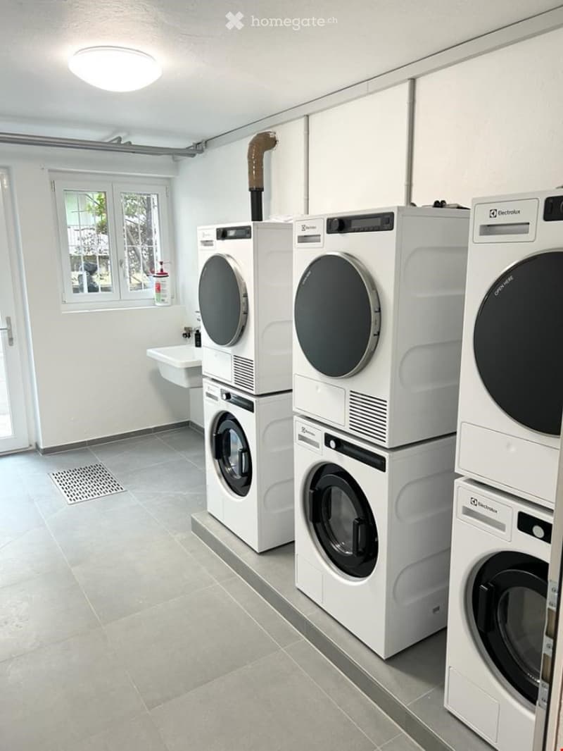 Clean, modern laundry room with multiple stacked washing machines and dryers, a small sink, and a window providing natural light.