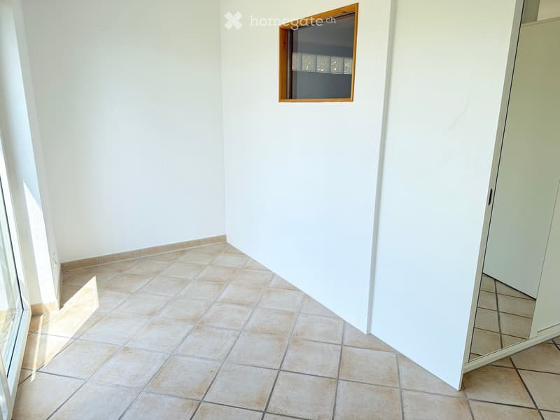 Empty room with tiled floor and white walls, featuring a window with wooden frame and a mirrored cabinet.