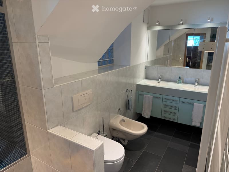 Modern bathroom with a toilet, bidet, double sink, large mirror, and light blue cabinets. The walls are tiled in light gray, and the floor is dark gray.
