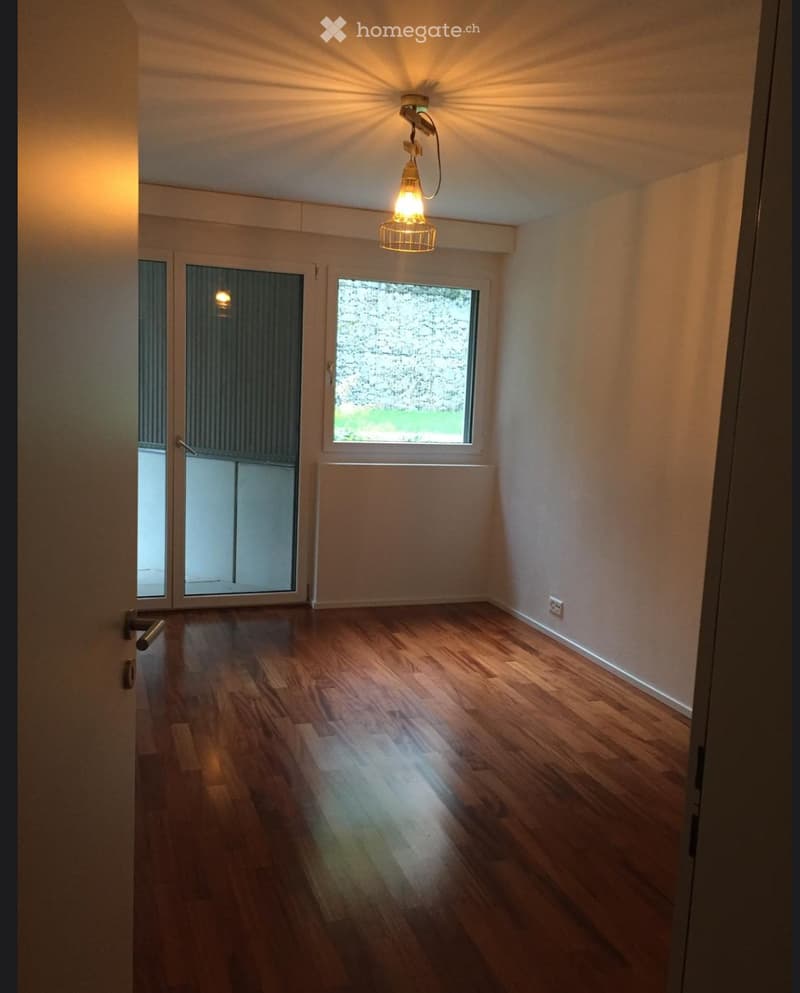 Empty room with wooden flooring, white walls, and large windows allowing natural light. Features a hanging light fixture and a door leading to another space.