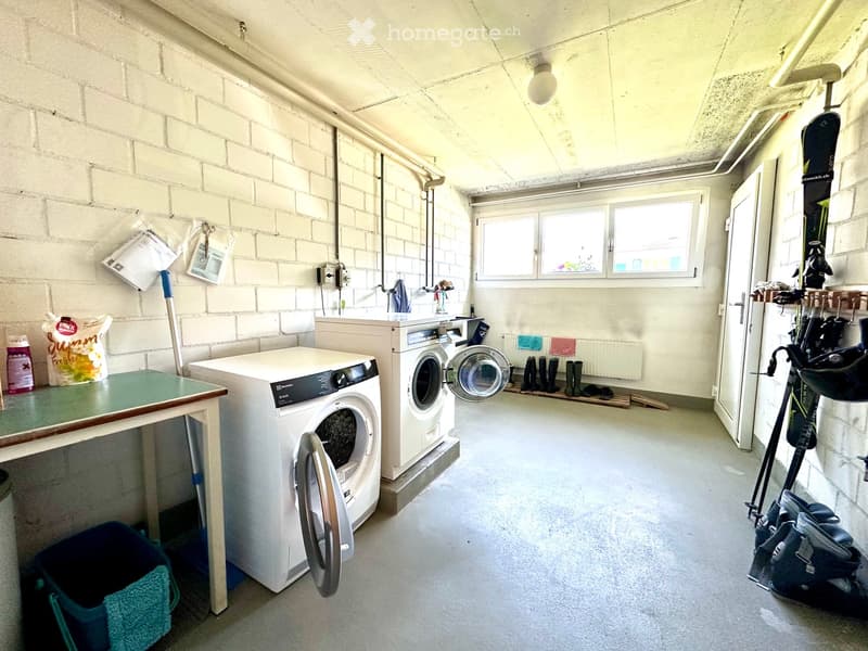 A small, well-lit laundry room with exposed brick walls, a washing machine, dryer, a small table, and various storage items including a backpack, snowboard, skis, and boots.
