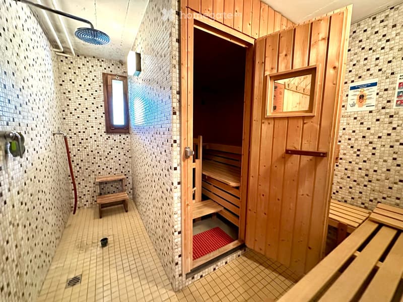 Bathroom with a shower area, wooden sauna bench, and a small window. The walls are tiled, and there is a wooden sauna door visible.