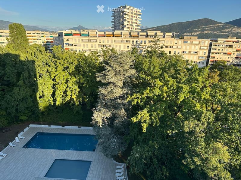 Aerial view of a rooftop garden with two swimming pools, surrounded by lush green trees and several lounge chairs, with apartment buildings and mountains in the background.