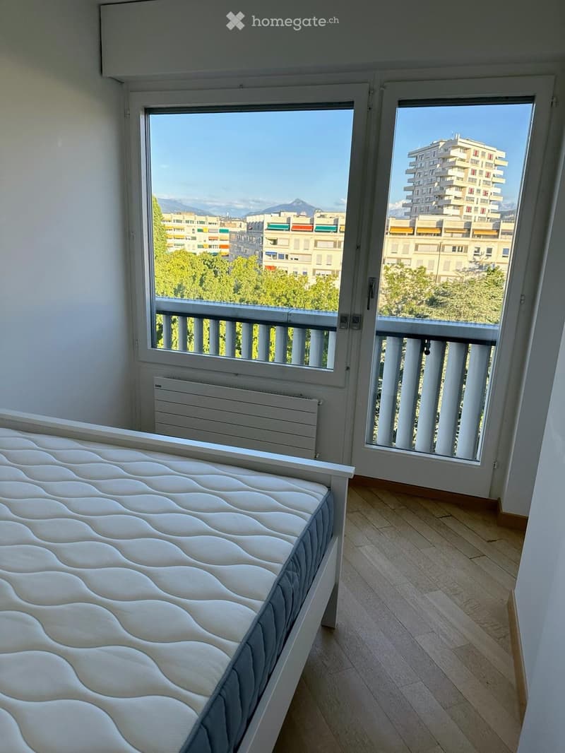 Bright bedroom with a white bed, wooden flooring, large windows offering a view of the city and mountains, and a radiator under one window.