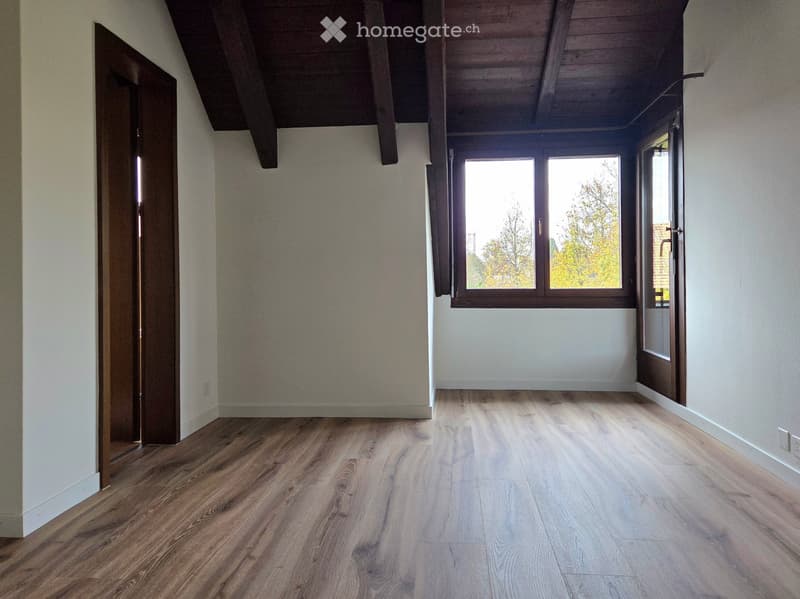 Empty room with wooden flooring, white walls, and large windows letting in natural light. The room features exposed wooden beams on the ceiling and a partially open door leading to another space.