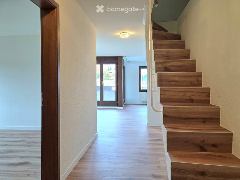Wooden staircase with a modern design, light-colored wooden flooring, and a recessed ceiling light. The hallway leads to a glass door with a view of the outside.