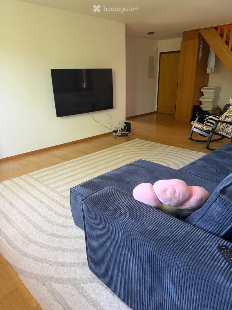 Living room with a blue sofa, pink pillows, a television mounted on the wall, a rocking chair, and a striped rug on a wooden floor.