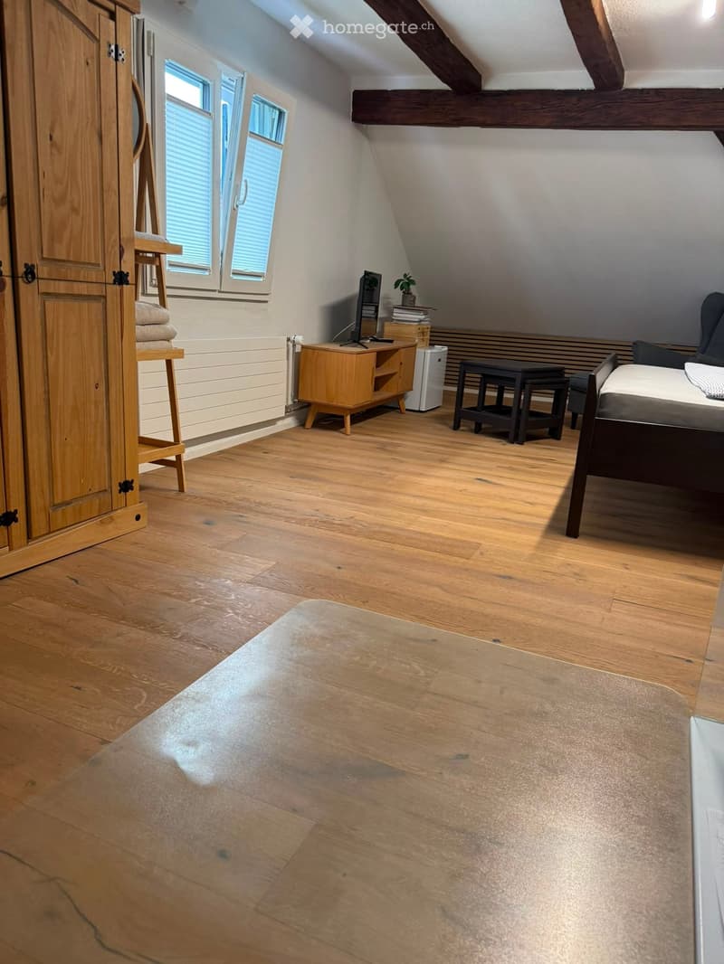Cozy attic room with wooden floors, slanted ceilings, and exposed beams. Features a sofa, small table, TV, and a few stools.