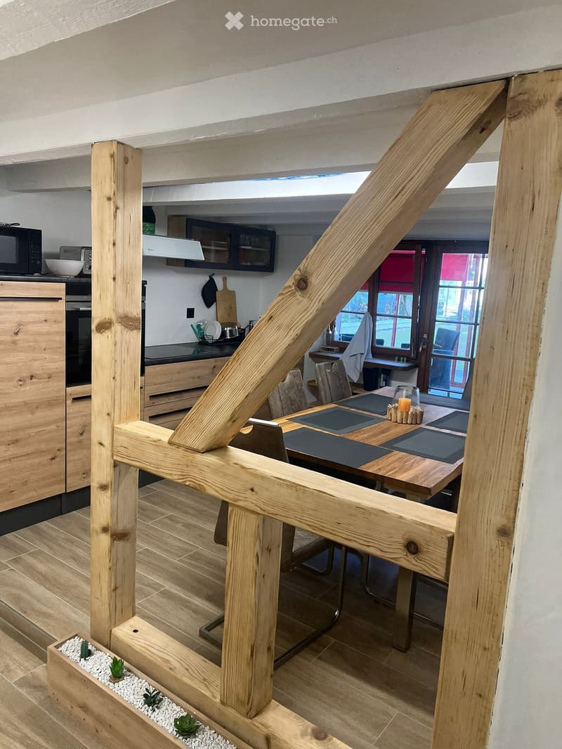 A modern, open-plan living and dining area with wooden beams, a large wooden dining table with chairs, a kitchen area with cabinets, microwave, and oven, and a small plant in a wooden planter.